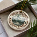A decorative ornament in a box, featuring a mountain landscape and pine trees, with the text "ROCKY MOUNTAIN NATIONAL PARK USA" printed on it.
