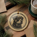 A ceramic ornament featuring a black and white illustration of a dog's face is displayed on a wooden surface, accompanied by a cup of coffee and pine branches.