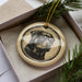 A ceramic ornament in the shape of a dog's head, featuring a black and white illustration, is displayed in a box with pine branches.