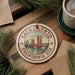 A ceramic ornament with the text "Sacramento, California USA" and an illustration of the Sacramento Bridge, placed on a wooden surface alongside a cup of coffee and some pine branches.