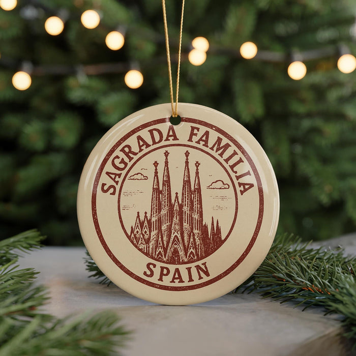 A circular ornament with the text "SAGRADA FAMILIA SPAIN" and an illustration of the Sagrada Familia church in Barcelona, Spain, is displayed in front of a blurred Christmas tree with twinkling lights.