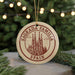 A circular ornament with the text "SAGRADA FAMILIA SPAIN" and an illustration of the Sagrada Familia church in Barcelona, Spain, is displayed in front of a blurred Christmas tree with twinkling lights.