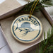 A round ornament with a blue and white design featuring a sailfish, placed inside a box with pine branches.
