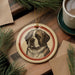A round ornament featuring a dog's face is displayed on a wooden surface, surrounded by pine branches and a cup of coffee.