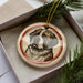 A round ornament featuring a dog's face is displayed in a box, surrounded by pine branches.