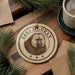A circular ornament featuring a monkey's face is displayed on a wooden surface, surrounded by pine branches and a cup of coffee.