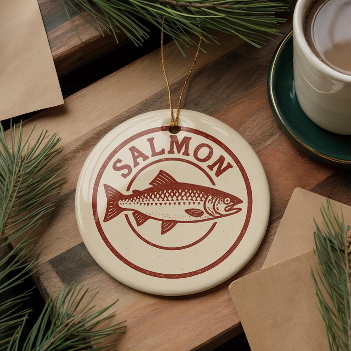 a round ornament with a salmon design, placed on a wooden surface next to a cup of coffee and some pine branches.