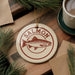 a round ornament with a salmon design, placed on a wooden surface next to a cup of coffee and some pine branches.