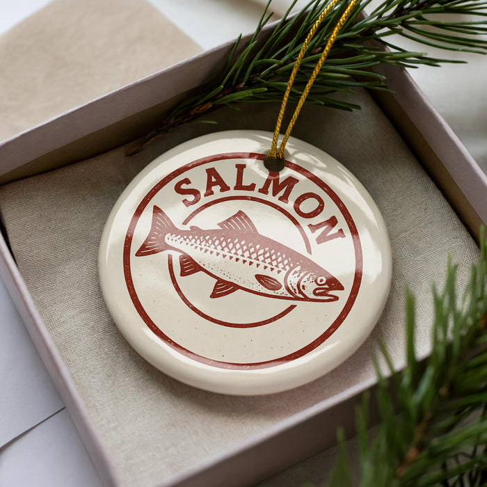 A white ceramic ornament with a red and brown design featuring a fish, placed inside a cardboard box with pine branches.