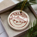 A white ceramic ornament with a red and brown design featuring a fish, placed inside a cardboard box with pine branches.