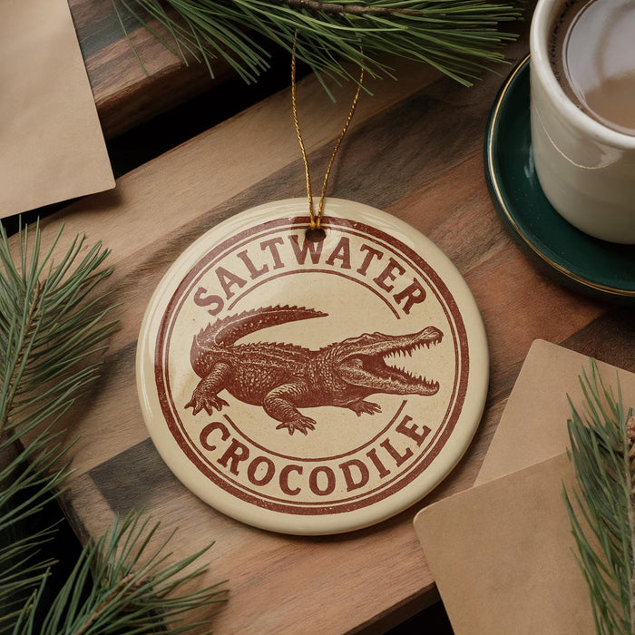 A round ornament featuring a crocodile is displayed on a wooden surface, surrounded by pine branches and a cup of coffee.
