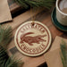 A round ornament featuring a crocodile is displayed on a wooden surface, surrounded by pine branches and a cup of coffee.
