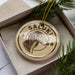 A circular ornament with a dog's head and the word "SALUKI" printed on it, placed inside a box with pine branches.