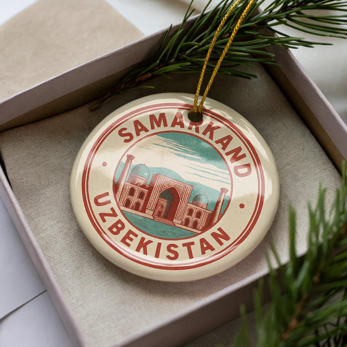 A circular ornament with the text "SAMARKAND UZBEKISTAN" printed on it, surrounded by a border of red and green colors. The ornament is placed inside a box, with a sprig of pine branches visible to the right side of the image.
