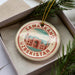 A circular ornament with the text "SAMARKAND UZBEKISTAN" printed on it, surrounded by a border of red and green colors. The ornament is placed inside a box, with a sprig of pine branches visible to the right side of the image.