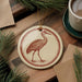 a round ornament with a bird illustration, placed on a wooden surface next to a cup of coffee and some pine branches.