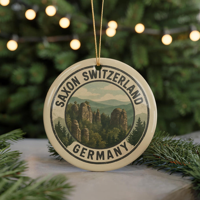 A round ornament with the text "SAXON SWITZERLAND GERMANY" and a scenic mountain landscape printed on it, hanging from a string.