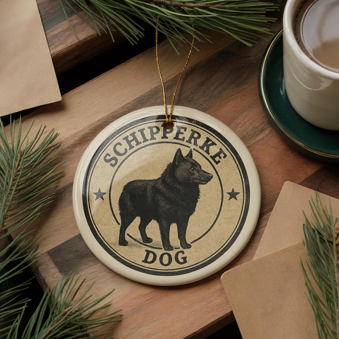 A round ornament featuring a black dog, with the text "SCHIPPERKE DOG" printed on it, is displayed on a wooden surface alongside a cup of coffee and some pine branches.