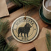 A round ornament featuring a black dog, with the text "SCHIPPERKE DOG" printed on it, is displayed on a wooden surface alongside a cup of coffee and some pine branches.