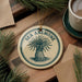 a circular ornament with the text "SEA ANEMONE" on it, surrounded by pine branches and a cup of coffee on a wooden surface.