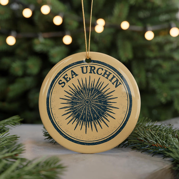A round ornament with the text "SEA URCIN" and a graphic of a sea urchin is displayed on a table, surrounded by pine branches and twinkling lights.