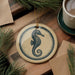a round ornament with a seahorse design, placed on a wooden surface next to a cup of coffee and some pine branches.
