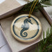 A round ornament featuring a seahorse design is displayed in a box, surrounded by pine branches.