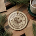 A round ornament featuring a dog's head and the text "SEALYHAM TERRIER" is displayed on a wooden surface, surrounded by pine branches and a cup of coffee.