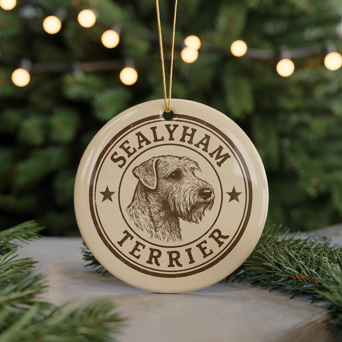 A round ornament featuring a terrier dog's head, with the text "SEALYHAM TERRIER" printed on it, hanging in front of a Christmas tree with twinkling lights.