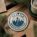A decorative ornament featuring the Seattle skyline, with the text "SEATTLE WASHINGTON" prominently displayed. The ornament is hanging from a string, and there are pine branches and a cup of coffee visible in the background.