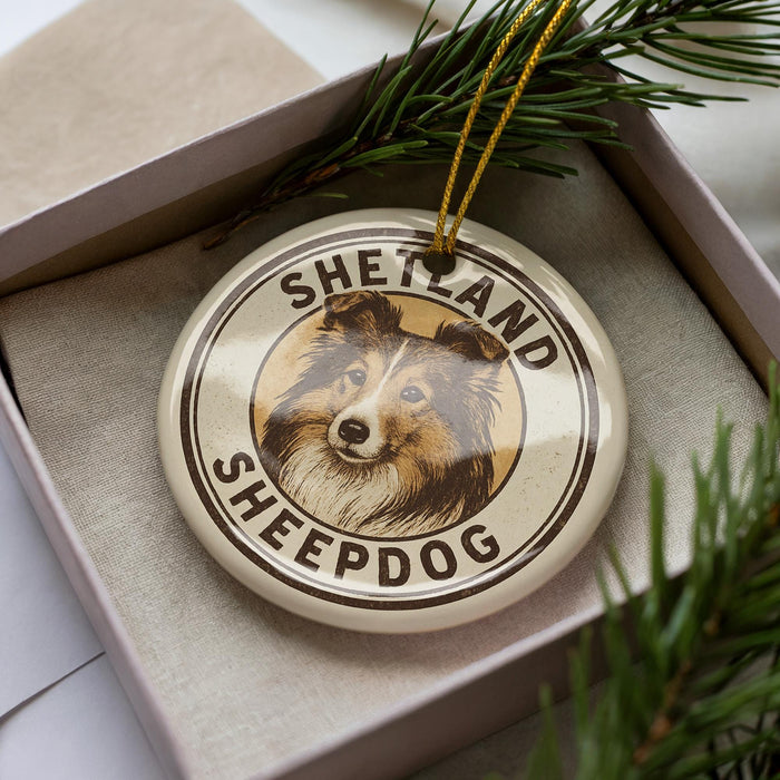A circular ornament with a picture of a dog's face and the text "SHELAND SHEEPPDOG" on it, placed in a box with pine branches.