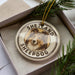 A circular ornament with a picture of a dog's face and the text "SHELAND SHEEPPDOG" on it, placed in a box with pine branches.