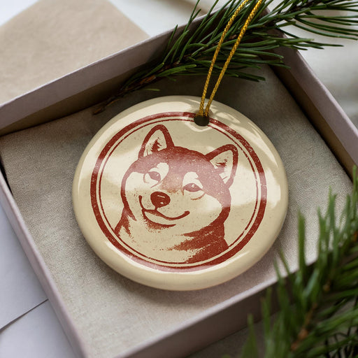 A ceramic ornament in the shape of a dog's head, with a red and beige color scheme, is displayed in a box surrounded by pine branches.