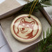 A ceramic ornament in the shape of a dog's head, with a red and beige color scheme, is displayed in a box surrounded by pine branches.