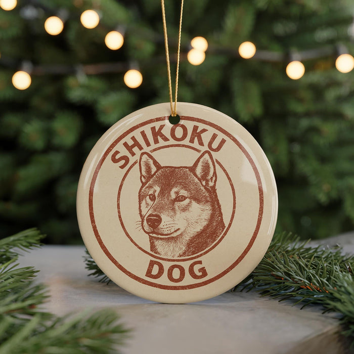 A circular ornament featuring a dog's head and the text "SHIKOKU DOG" in a brown color scheme, hanging from a string against a blurred background of a Christmas tree.