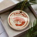 A round ornament with a shrimp design, placed inside a box with pine branches.