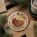 a circular ornament with the text "SIMIEN MOUNTAINS ETHIOPIA" on it, surrounded by pine branches and a cup of coffee on a wooden surface.