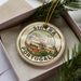 A circular ornament with a picture of a building and the text "SINCTRA PORTUGAL" on it, placed on a box with pine branches.