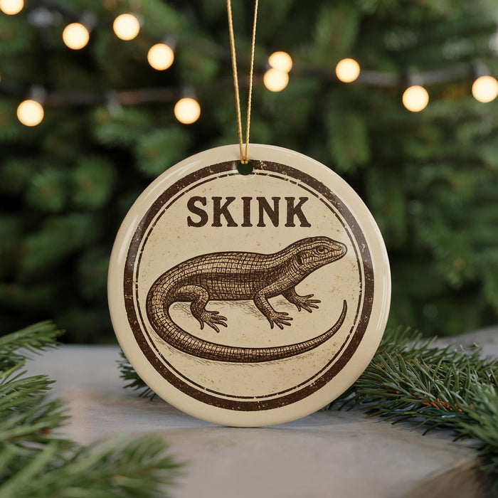 A round ornament with a lizard image and the word "SKINK" written on it, hanging in front of a blurred Christmas tree with lights.