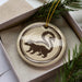 A ceramic ornament in the shape of a skunk, with a brown and white design, is displayed in a box with pine branches.