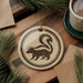 a round ornament with a black and white illustration of a skunk, hanging on a wooden surface. There is also a cup of coffee and some pine branches in the background.