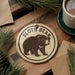 A circular ornament featuring a sloth bear illustration is displayed on a wooden surface, accompanied by a cup of coffee and pine branches.