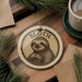 A round ornament featuring a sloth, a type of slow-moving mammal, is displayed on a wooden surface alongside a cup of coffee and some pine branches.