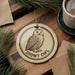 A round ornament featuring a snowy owl is displayed on a wooden surface, surrounded by pine branches and a cup of coffee.
