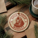 A round ornament with a monkey image and the text "SNUB-NOSED MONKEY" is displayed on a wooden surface, surrounded by pine branches and a cup of coffee.