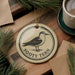 A round ornament featuring a bird, specifically a seagull, is displayed on a wooden surface alongside a cup of coffee and some pine branches.