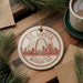 A decorative ornament featuring the St. Louis, Missouri skyline, with the text "St. Louis Missouri - USA" printed on it. The ornament is hanging from a string and is placed on a wooden surface, surrounded by pine branches and a cup of coffee.