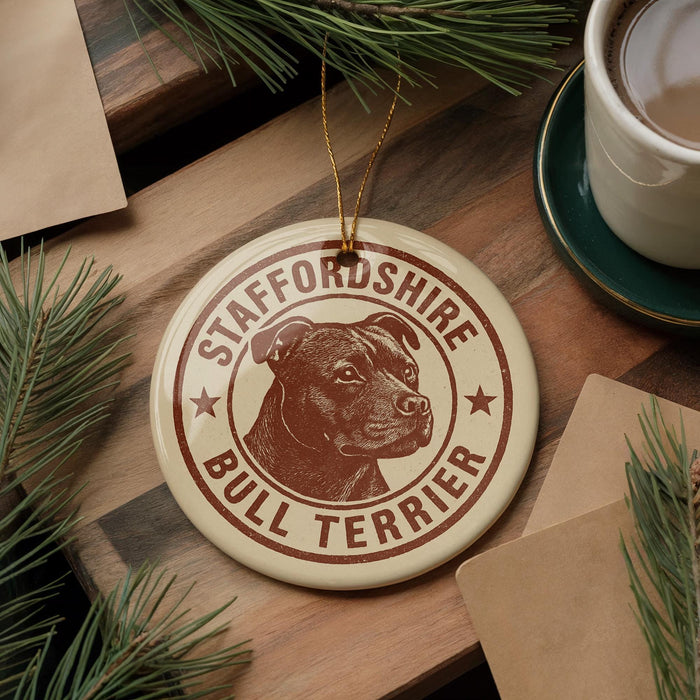 A circular ornament featuring a bull terrier dog's head, with the text "STAFFORDSHIRE BULL TERRIER" printed on it, is displayed on a wooden surface alongside a cup of coffee and some pine branches.