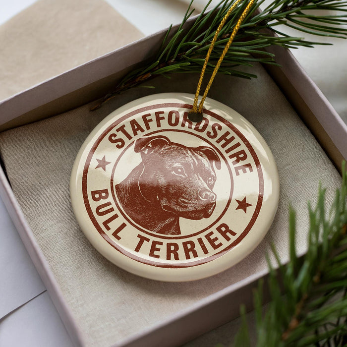 A circular ornament with a bull terrier image and the text "STAFFORDSHIRE BULL TERRIER" printed on it, placed inside a box with pine branches.