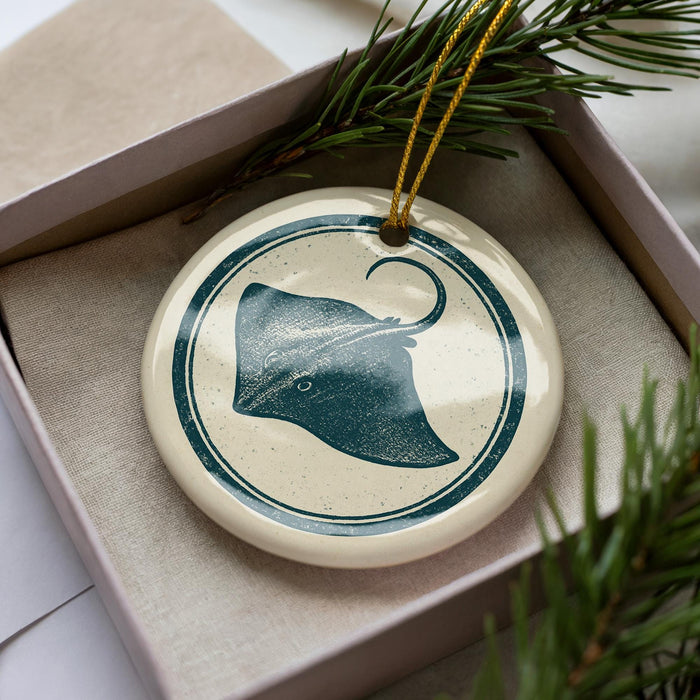 A ceramic ornament in the shape of a stingray is displayed in a box, surrounded by pine branches.